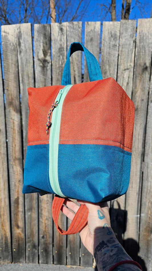 Slightly smaller than the Howdy OG this Orange and Teal poly-coated canvas zipper bag with a seafoam green zipper. This bag has two long handles on either end so you can hang it, or loop it around your handlebars!