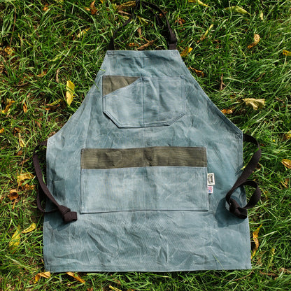 Blue and grey apron with black straps. Hand Waxed and laying in the grass.
